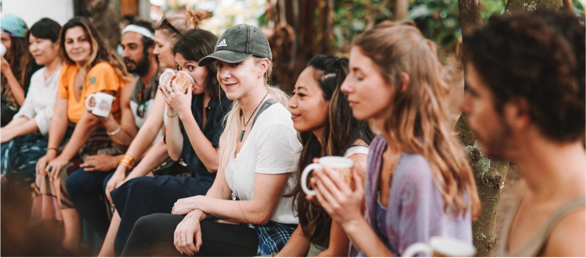 A group of young people sitting outdoors on a bench, holding white ceramic mugs. The scene is softly blurred with warm natural light filtering through trees, and no clear product brand is visible.